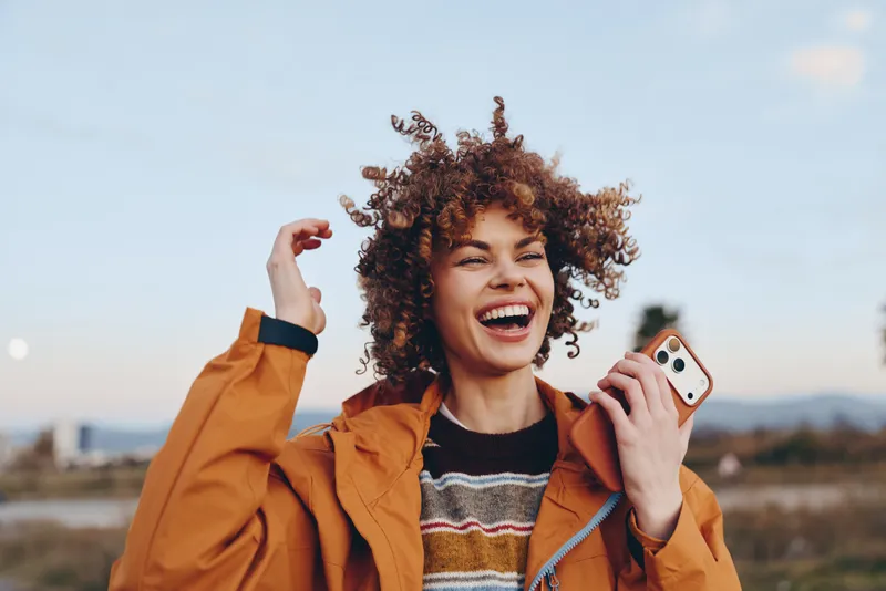 Smiling woman with curly hair outdoors in an orange jacket. She shows laughter while holding a smartphone, lifestyle moment with soft bokeh background and warm natural light cheerful vibe.