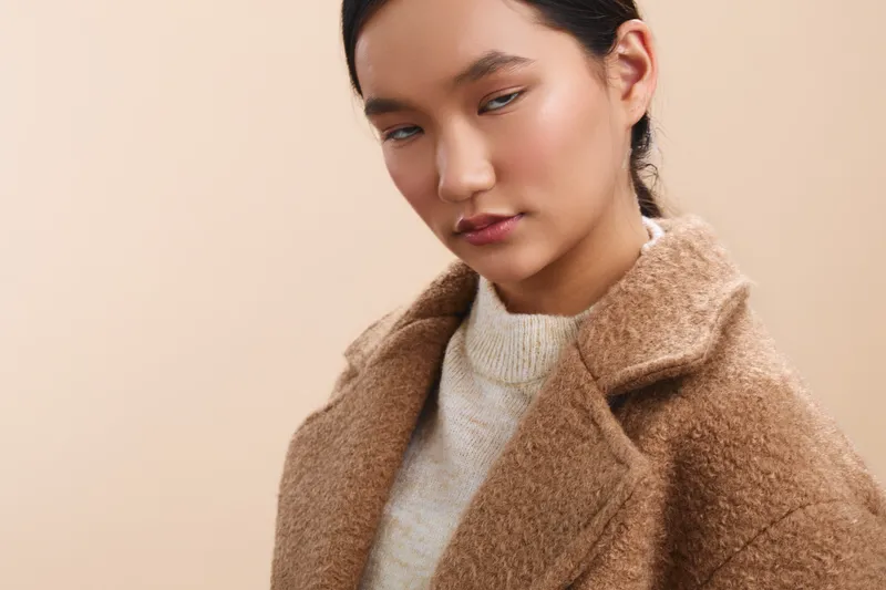 asian woman wearing a tan wool coat and light sweater stands in a minimal studio, looking thoughtful and poised, conveying calm confidence and modern style.