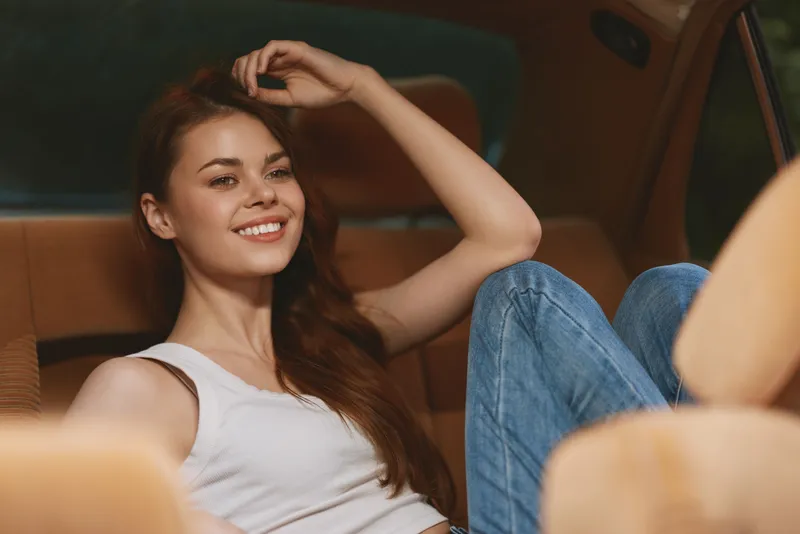 Young woman with long hair sitting in a car, smiling and relaxed, dressed in a white tank top and blue jeans, warm tones, concept of freedom and joy.