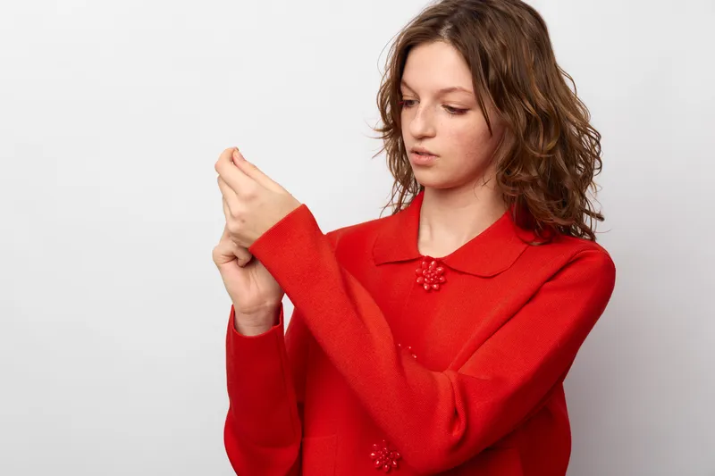 Young woman in red sweater looks at her wrist with concern, isolated on white background. Concept of pain, discomfort, and health awareness in everyday life.