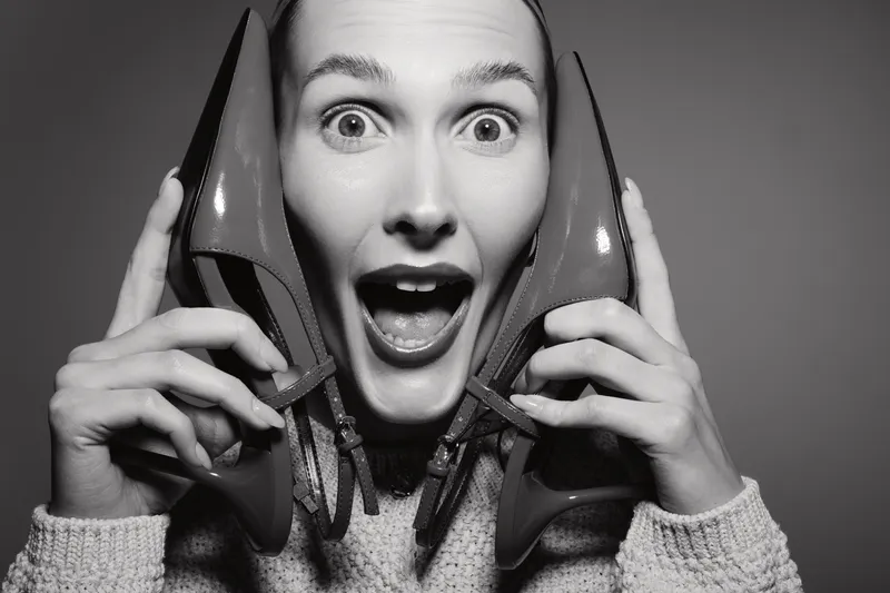 Surprised white woman with light skin holding red high heels near face, wearing knitted sweater in studio lighting.