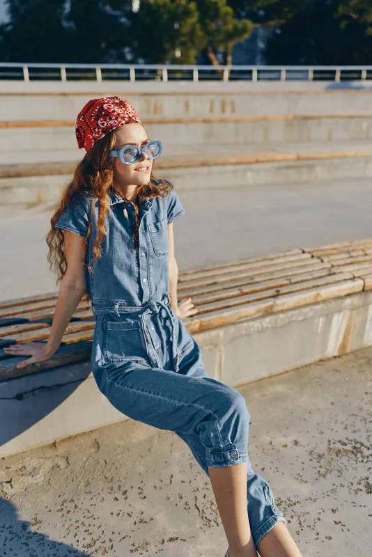 Lifestyle woman wearing modern boho-western denim jumpsuit and red bandana relaxing outdoors in warm sunlight with film color tones, enjoying casual street style fashion.