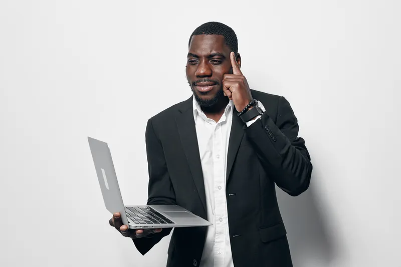 Black man with dark skin in business suit holding laptop and pointing to head for idea or thinking concept on white background. Professional African male with confident expression.