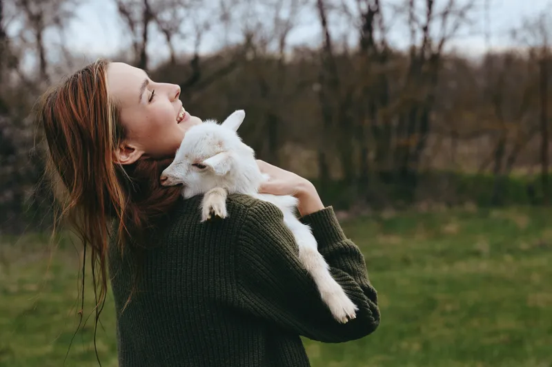 Cheerful woman outdoors, goat kid in her arms, affection and cuddling in a natural outdoor lifestyle, sunlit meadow and calm nature backdrop, candid and warm.