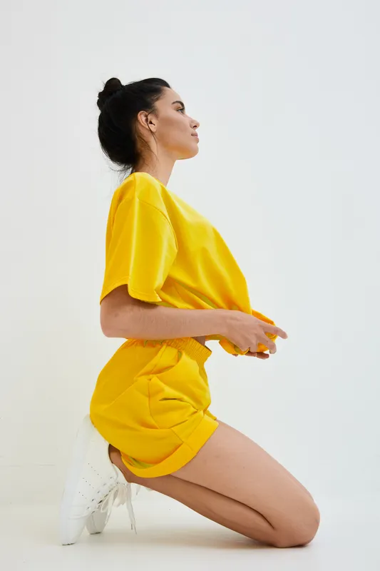 Young woman kneeling in casual yellow outfit and white sneakers against white background, displaying calm and focused expression with minimalistic style and bright colors.