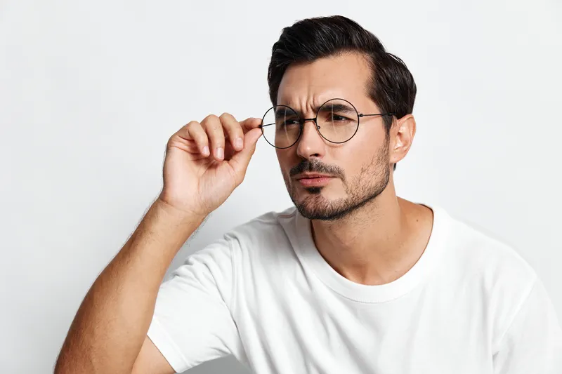 Thoughtful man wearing round glasses looking away while adjusting his eyewear with one hand, isolated on light solid background in a casual white t-shirt.