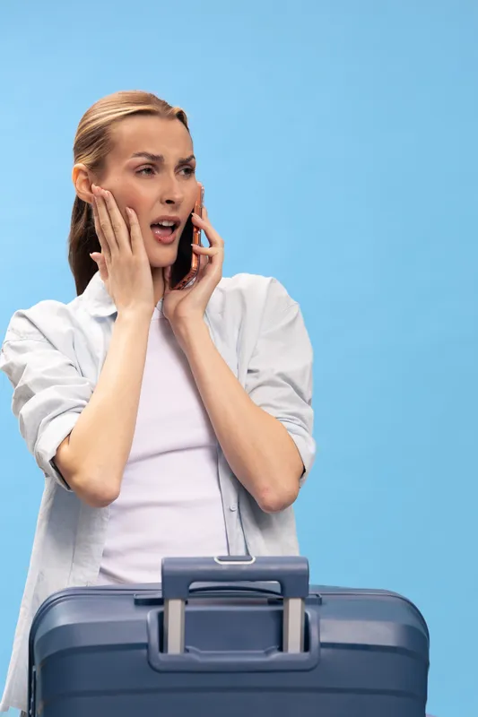 Worried woman talking on phone with anxious expression, holding face with hand near blue suitcase. Concept of travel stress, urgent communication, and unexpected problems during trip.