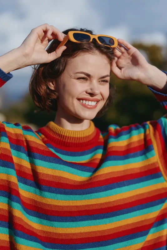 Woman smile portrait outdoors in colorful striped sweater and yellow sunglasses, happy young adult adjusting shades with joyful expression, natural light candid lifestyle moment.