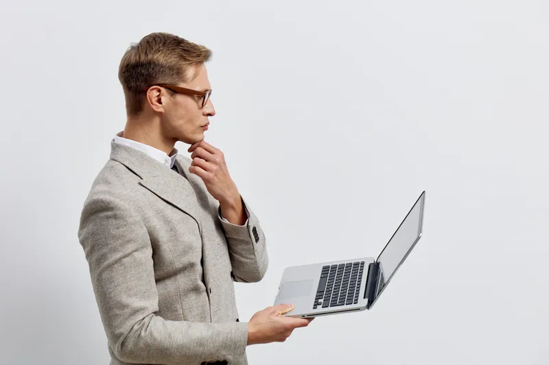 Professional young man in glasses and stylish gray blazer holding a laptop with serious expression, standing against plain white background, focused on work, technology concept, modern office style.