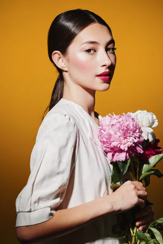Stylish woman with elegant makeup and smooth hair poses holding pink and white flowers against a warm orange background. Beauty, creative, fashion concept.