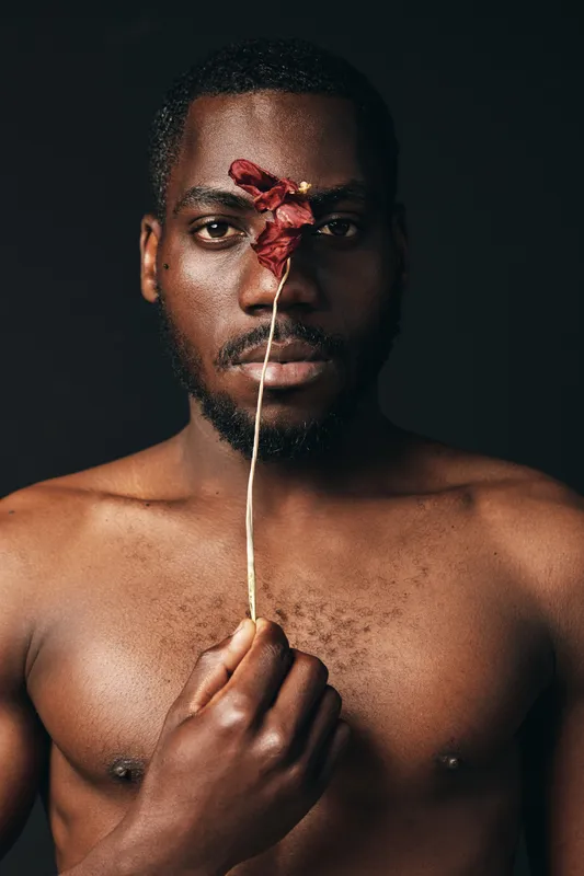 African man with dark skin holding dried red flower in front of face, creative conceptual portrait with visual style, bare chest and serious expression on dark background.