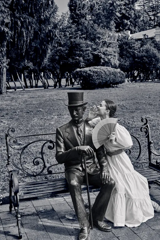 A woman in a vintage dress leans toward a bronze statue of a man with a top hat on a park bench, creating a romantic, nostalgic scene in a sunny day