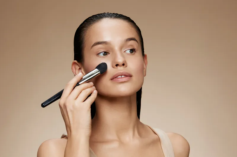 Young woman applying makeup with a brush on her cheek, showcasing a natural beauty look against a warm beige background, exuding confidence and elegance