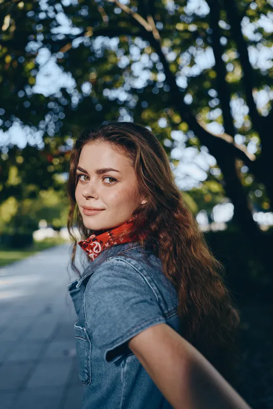 smiling woman with curly hair wearing denim jacket and red scarf stands outdoors under green leafy trees enjoying sunny day