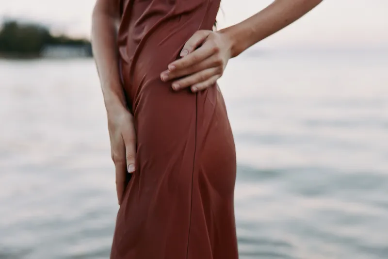 A close up of a person in a rust red dress standing by calm water, hand resting on hip, conveying elegance, confidence, and a relaxed coastal mood.