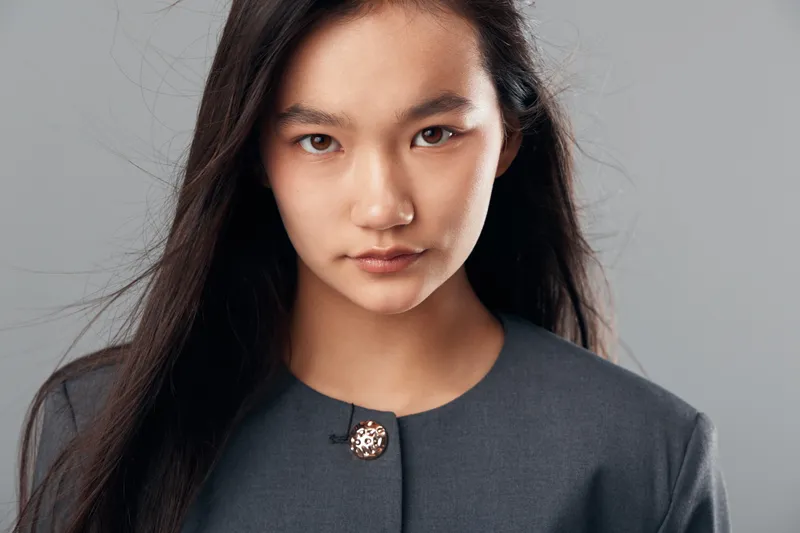 Portrait of young Asian woman with long black hair wearing gray professional jacket with brooch, looking confidently at camera in studio with simple gray background and natural light.
