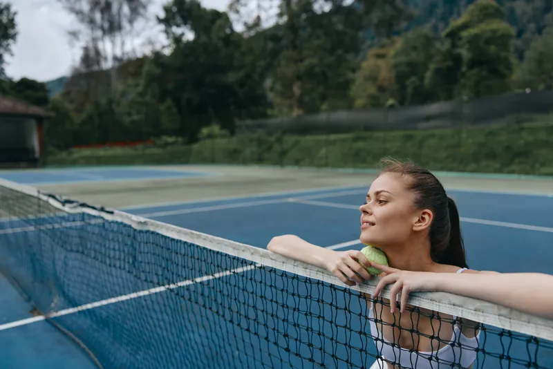 Young female tennis player relaxing by the net, wearing athletic gear, showcasing determination and joy on the vibrant blue court