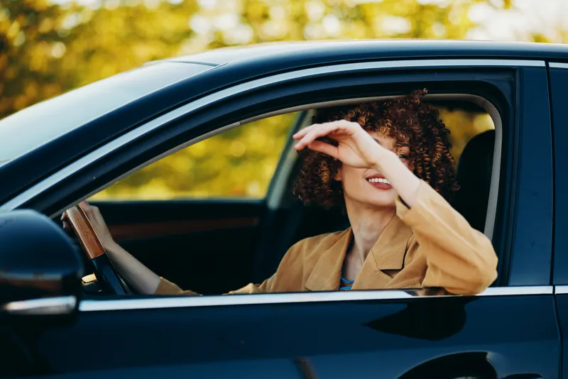 A driving woman with curly hair wearing a tan blazer sits in a car and raises her hand as she glances ahead, captured in a retro film color mood