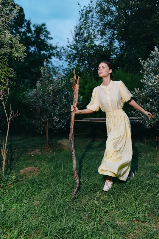 A woman in a pale yellow dress outdoors in a garden moves with motion, carefree and in summer, walking across the grassy area, holding a long branch.