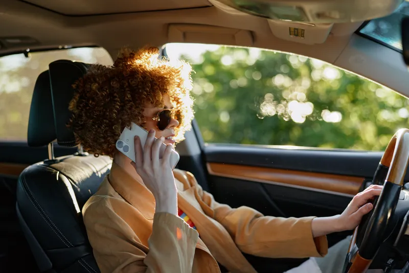 A woman with curly hair drives a car while talking on a phone, wearing a beige shirt. Sunlight fills the interior, creating a casual, everyday moment in a travel scene.