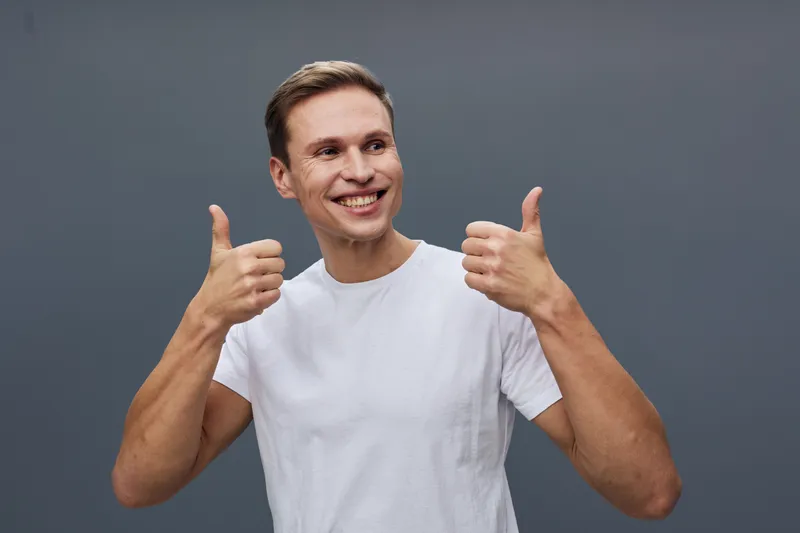 Smiling man wearing casual white t-shirt giving thumbs up gesture look at camera studio portrait on plain gray background people lifestyle concept