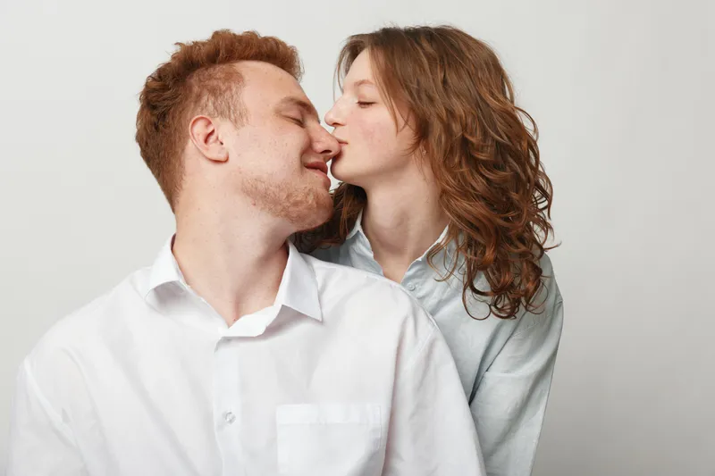 Young couple in casual clothes shares a tender moment with a gentle kiss on the nose. Intimate love and affection captured in a bright studio with a plain background.