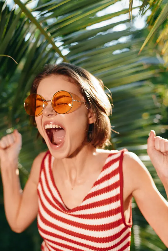 A joyful woman wearing a striped sleeveless top and round sunglasses cheers with raised fists in a sunny outdoor setting, radiating energy, confidence, and carefree optimism.