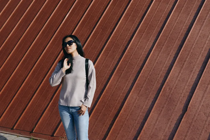 Woman wearing sunglasses, sweater and jeans stands beside a diagonal brown wall, showcasing casual street fashion, urban style and a relaxed travel vibe with a small backpack under sunlight