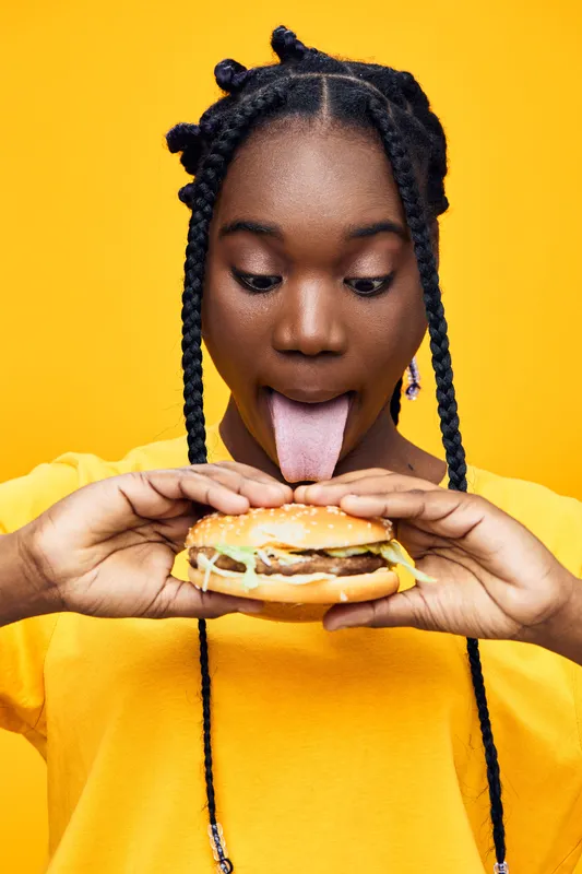 Happy young woman with braids holding a burger against a bright yellow background, expressing excitement and enthusiasm for delicious fast food