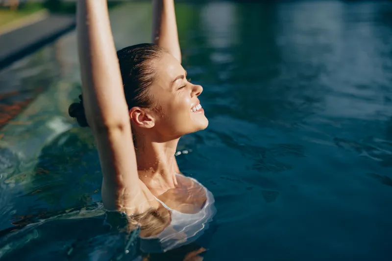 Smiling woman in a pool raises arms, enjoying sunlit water. Wellness lifestyle moment, relaxation evident in a bright poolside setting, with blue water and warm sunlight.