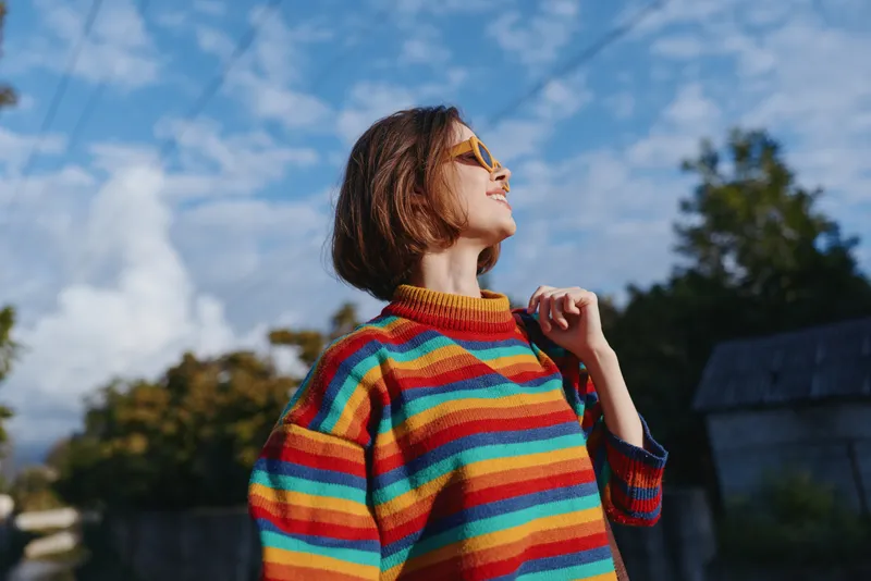Woman in a rainbow sweater and sunglasses looking up, short hair and smiling outdoor in a suburban street. Cozy striped knit, mid twenties model, lifestyle travel and fashion vibe.