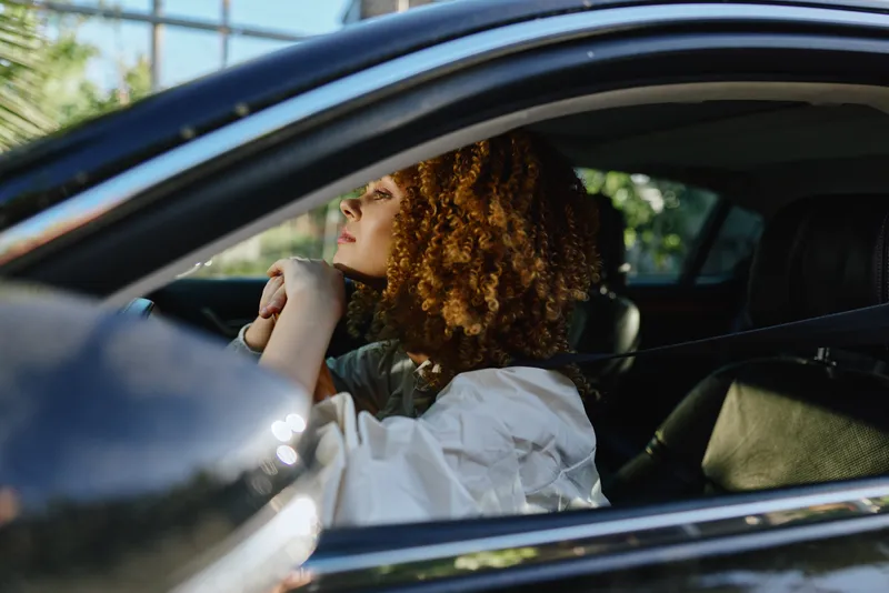 Woman with curly hair sitting inside a black car wearing a white blouse, looking outside thoughtfully in natural daylight with blurred green background.