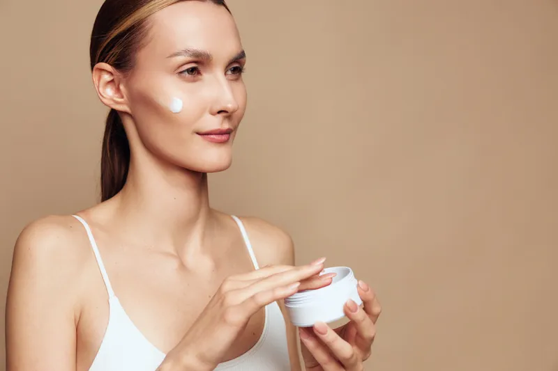 Young woman with clear skin applying white face cream on cheek. Close-up portrait on beige background. Concept of skincare, beauty routine, and natural wellness. Soft lighting highlights healthy skin.