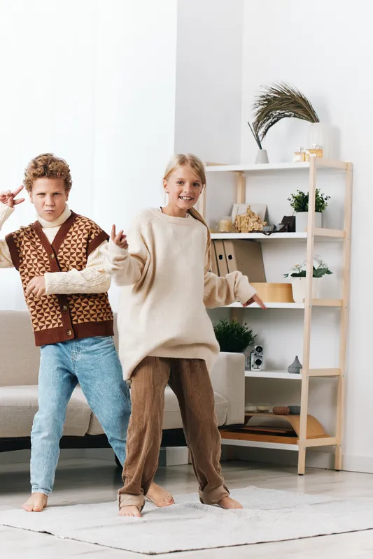 Two children dance playfully indoors, wearing cozy casual outfits as they move to music in a bright modern living room with a light rug, shelves, and decorative plants.