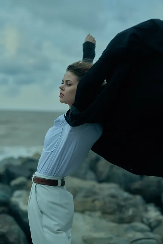 Windy beach scene shows a confident woman in a light blue shirt and white trousers, cape flying behind her as she strides forward, symbolizing leadership, determination and bold professional energy.