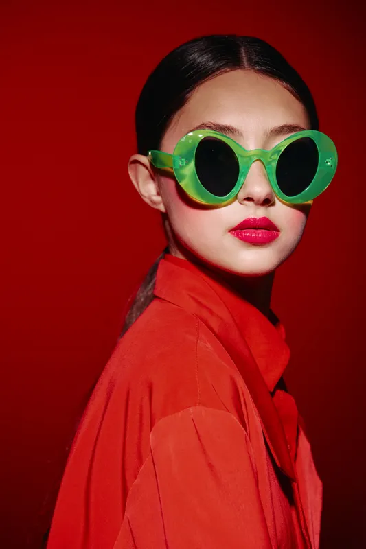 Young woman wearing oversized green sunglasses and vibrant red outfit poses against bold red background. Fashion beauty portrait capturing striking contrast and elegant style.