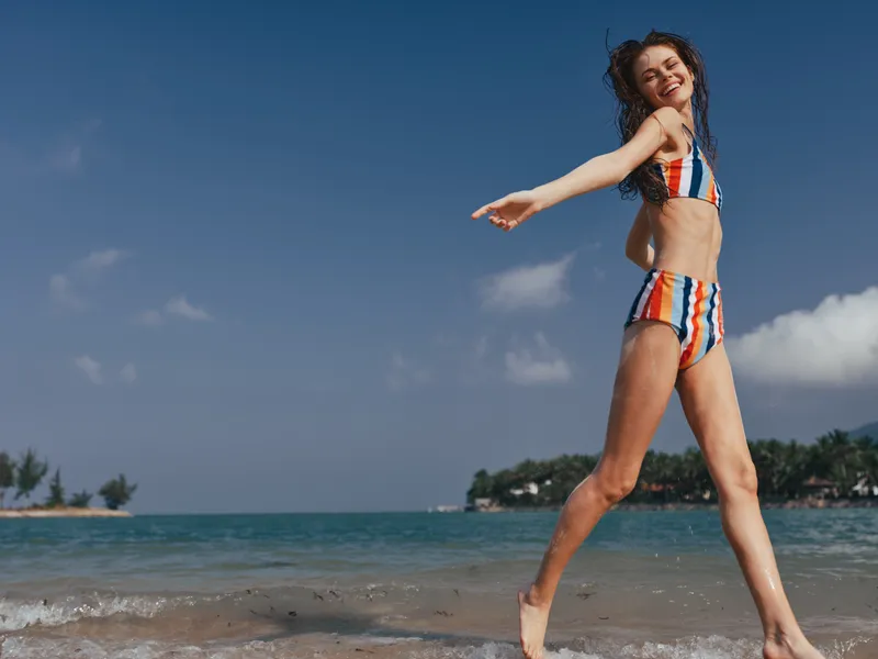 Young woman in a striped bikini runs along the sunny shoreline, arms extended, laughing as waves splash, capturing carefree summer energy and dynamic seaside motion.