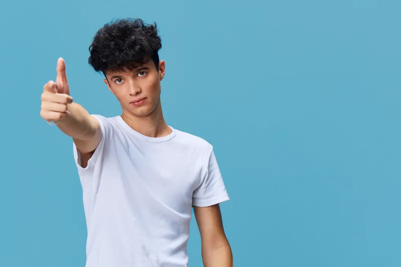 Young man in white t-shirt pointing at camera with confident expression against bright blue background, casual style and modern hairstyle.