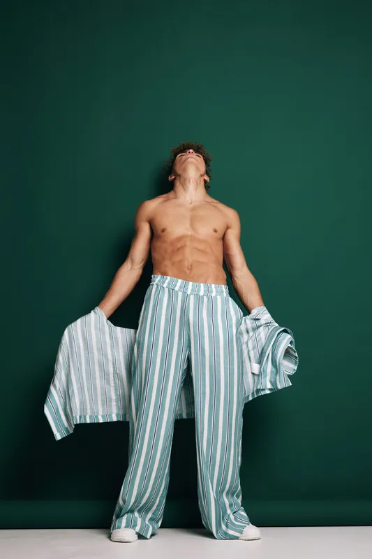 Shirtless man standing with striped pants and holding matching shirt, looking up against green background in casual fashion pose with relaxed expression.