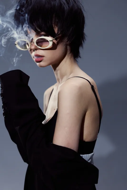 Stylish woman wearing edgy sunglasses and black jacket smoking with a moody gray background.