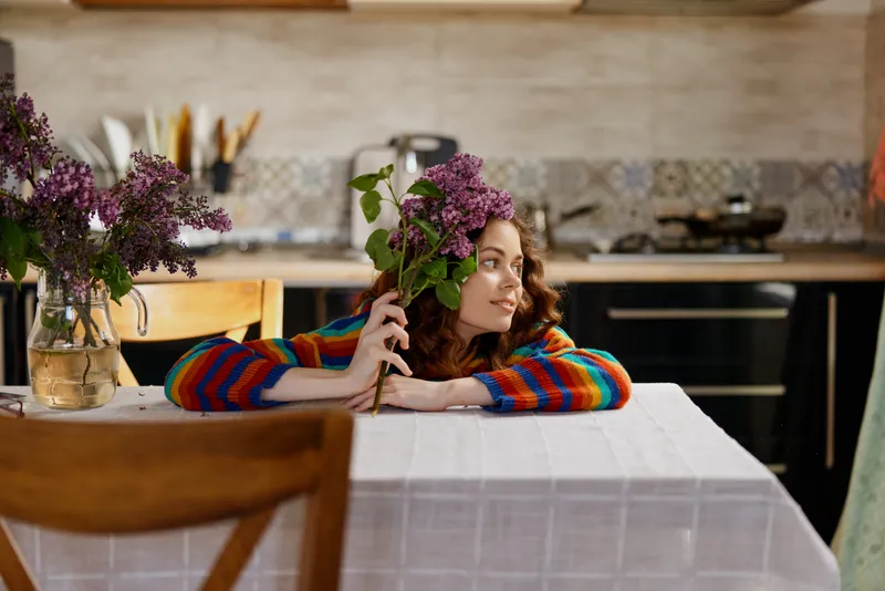 Smiling woman with wavy hair resting her chin on a flower bouquet at a dining table. Cozy kitchen backdrop, warm colors, casual home vibe, soft light creates a serene mood.
