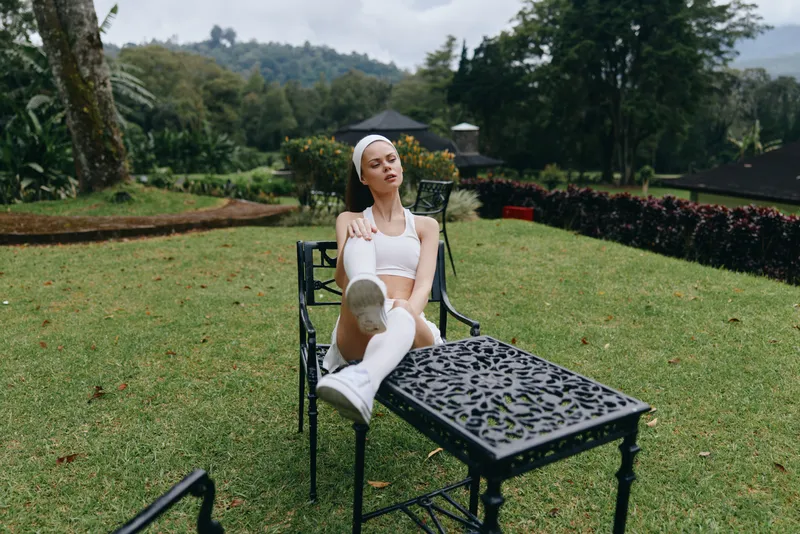 Young woman in sporty white outfit relaxing in a tranquil garden setting, reflecting peace and wellness through nature and fashion