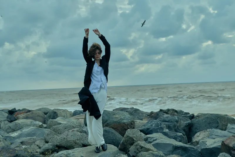 Woman on coastal rocks with arms raised, windy sea breeze, flowing coat, white shirt and trousers, seagull overhead, overcast sky, dramatic scene of resilience and freedom by the shore