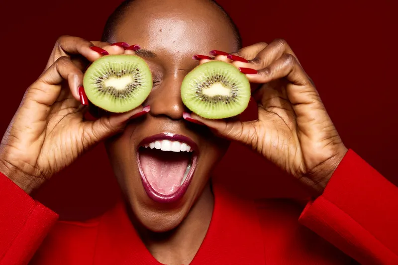 Happy African woman with dark skin wearing red holds two kiwi halves in front of her eyes, smiling widely on red background. Concept of healthy lifestyle, fruit, and joyful expression.