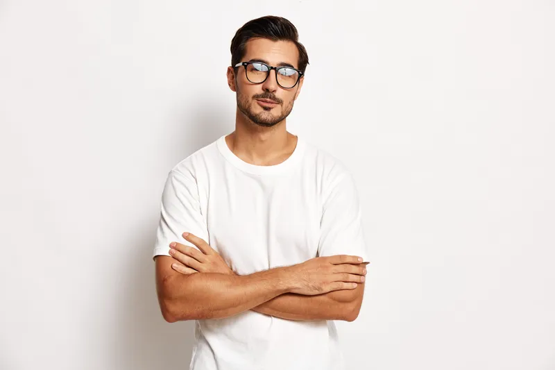 Confident young man with glasses wearing a white t-shirt stands with arms crossed, looking directly at the camera against a plain white background. Stylish portrait concept.