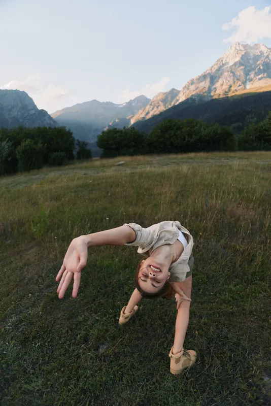 Young woman in a meadow outdoor scene performing a backbend, smiling at the camera, wind in hair, mountains in the background, colorful nature, relaxed lifestyle moment