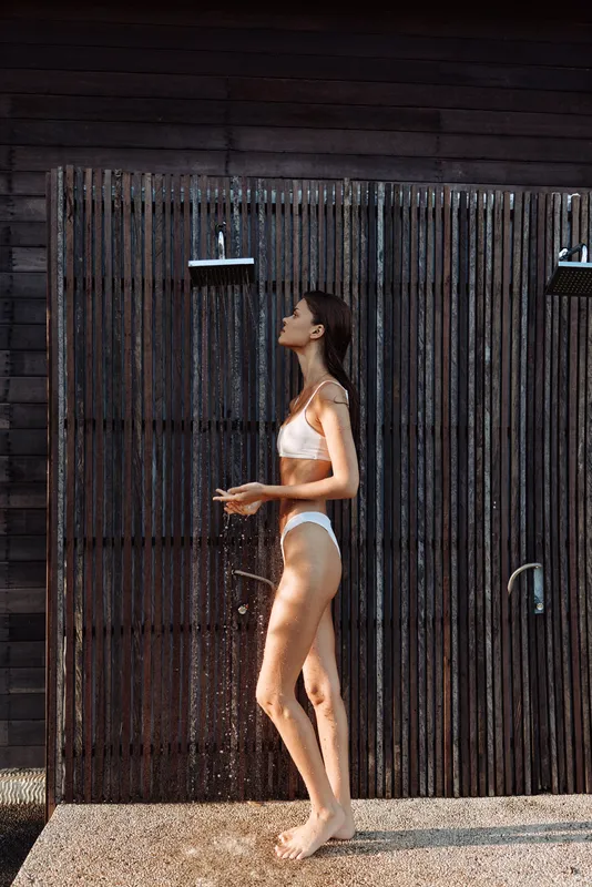 A young woman in a white bikini stands in an outdoor shower beside a weathered wooden fence, sunlit and relaxed, water droplets catching light