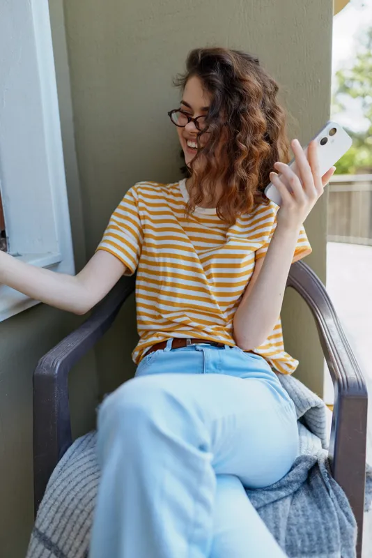 Casual woman relaxes on balcony chair, wearing striped yellow t shirt and light jeans, browsing smartphone, joyful mood, home lifestyle, candid moment, natural light, modern setting
