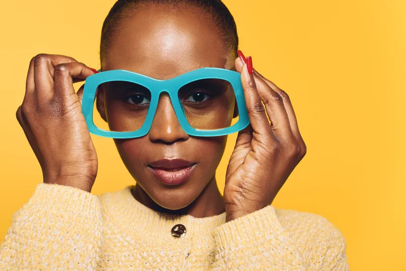 Close-up of confident dark skinned African woman wearing large blue glasses and yellow sweater on yellow background. Concept of fashion, style, beauty, and self-expression.