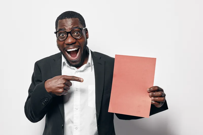 African American man with dark skin wearing glasses and suit holding blank paper and pointing at it with excited expression on white background for presentation or advertisement.